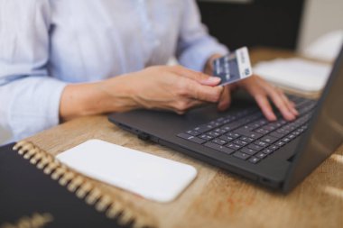 İş kadınının evde laptopu ve kredi kartıyla alışveriş yaparken çekilmiş görüntüsü mutfak masasında.