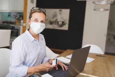 Genç ve güzel bir iş kadını dizüstü bilgisayarı ve kredi kartıyla mutfak masasında alışveriş yapıyor.