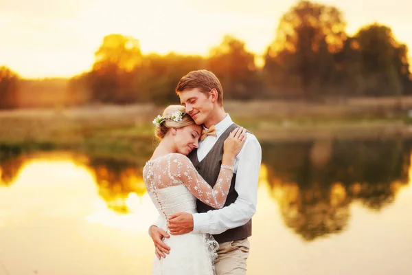 La novia y el novio están junto al lago, después de la ceremonia de la boda. Los recién casados sonríen, son felices. Foto en teñido cálido . — Foto de Stock