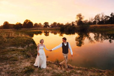 Gelin ve damat kırmızı günbatımı göl çerçevede akşamları yürümek. Genel planı