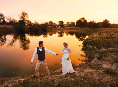 Gelin ve damat kırmızı günbatımı göl çerçevede akşamları yürümek. Genel planı