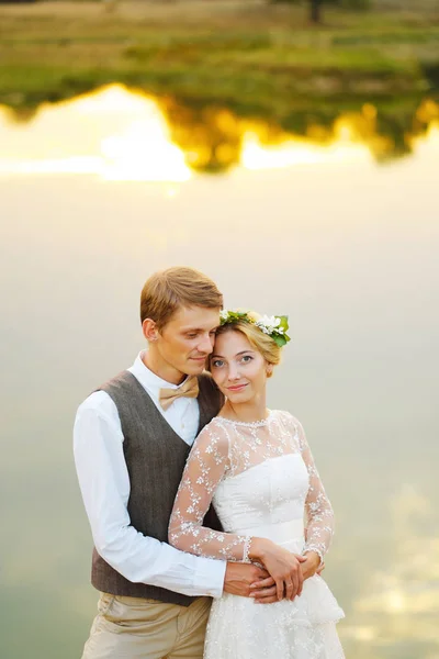Bruden och brudgummen kramas mot en sjö. Vattnet reflekterar molnen — Stockfoto