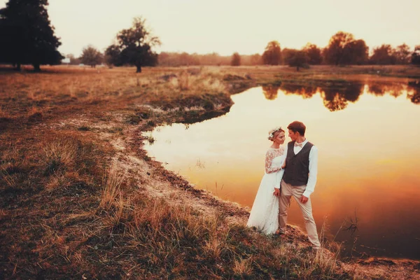 Bruden och brudgummen poserar mot bakgrund av sjön i röd solnedgång. Övergripande plan — Stockfoto
