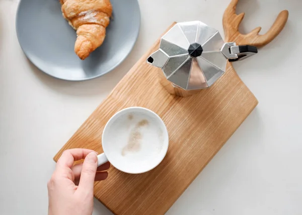 Xícara de chá na mão, croissant e cafeteira na mesa — Fotografia de Stock