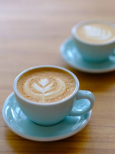 Två kopp kaffe i en vacker blå kopp på ett träbord i ett kafé. En cappuccino mugg med mönster på skummet. — Stockfoto