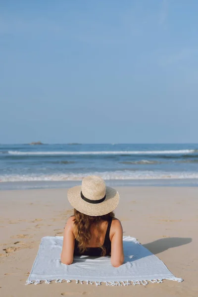 Achteraanzicht. Meisje in een stro hoed kijkt naar de oceaan zonnen op het zand — Stockfoto