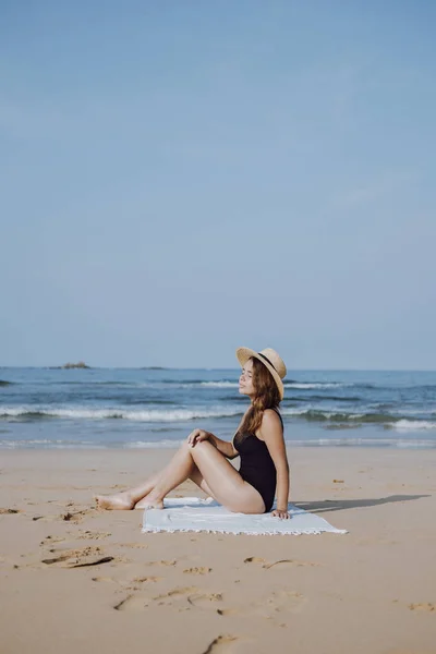 Meisje in een stro hoed zittend op het strand in de buurt van de Oceaan — Stockfoto
