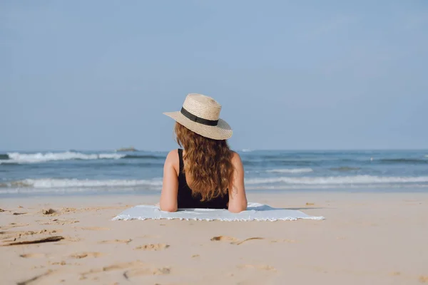 Achteraanzicht. Meisje in een stro hoed kijkt naar de oceaan zonnen op het zand — Stockfoto