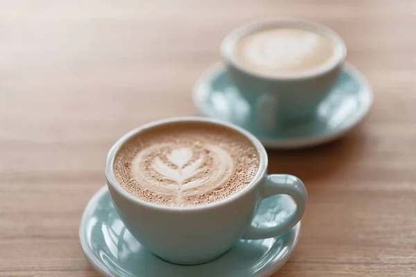 Två kopp kaffe i en vacker blå kopp på ett träbord i ett kafé. En cappuccino mugg med mönster på skummet. — Stockfoto