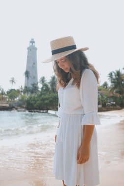 Bir deniz feneri önünde sahilde şapka içinde genç bir kadın duruyor