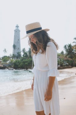 Bir deniz feneri önünde sahilde şapka içinde genç bir kadın duruyor