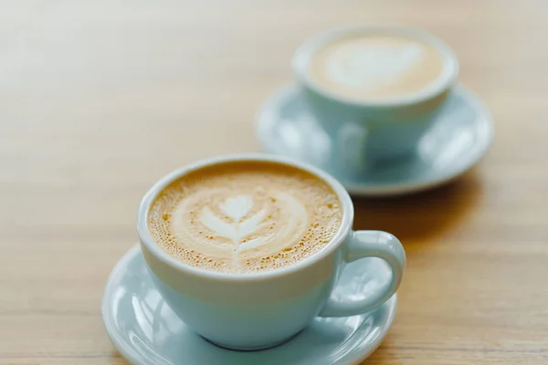 Två kopp kaffe i en vacker blå kopp på ett träbord i ett kafé. En cappuccino mugg med mönster på skummet. — Stockfoto