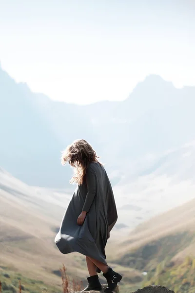 Joven hermosa chica en un vestido largo está de pie junto al acantilado en el fondo de las montañas — Foto de Stock