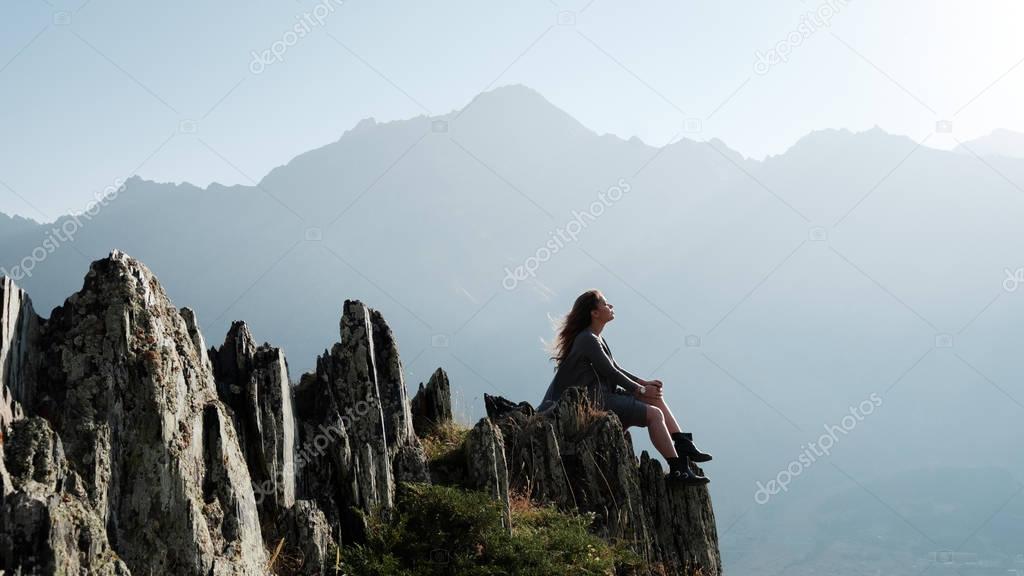 A beautiful girl in a flying dress  sits on a cliff overlooking the mountains