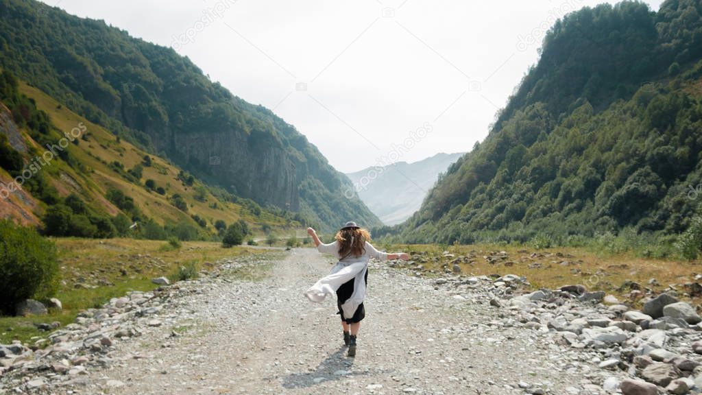 A girl in a hat walks along the middle of the road towards the mountains 