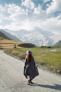 Güzel bir kız bir uçan elbise ve şapka dağlara doğru yol boyunca gider