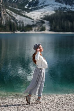 A girl in a hat and a long dress is walking along the lake. Lago di Bries in the north of Italy clipart