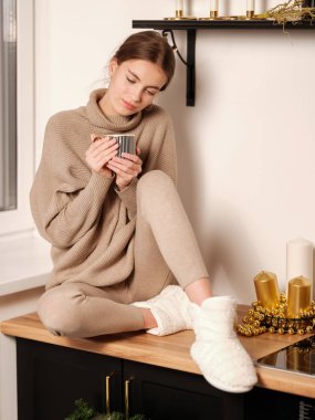 Elinde çay fincanı tutan ve mutfaktaki ahşap masada oturan rahat ev kıyafetleri içinde genç ve çekici bir kadın. 