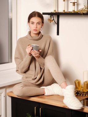 Elinde çay fincanı tutan ve mutfaktaki ahşap masada oturan rahat ev kıyafetleri içinde genç ve çekici bir kadın. 