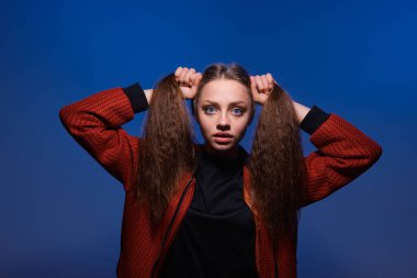 Saçlarını elleriyle atkuyruğu yapan, stüdyoda mavi arka planda poz veren sevimli bir genç kız..