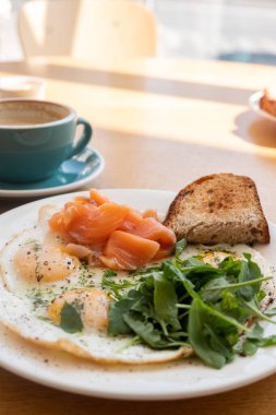 Kızarmış yumurtalı tabak ve ekmekle bitkiler ve ahşap masada kapuçino bardaklı balık.