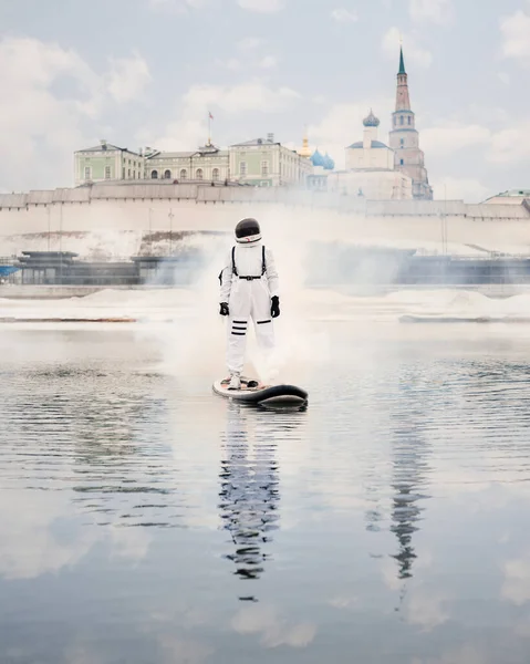 Astronaut ve skafandru stojí na sup prkně a plave na řece ve velkém městě. — Stock fotografie