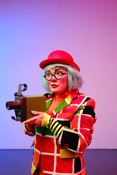 Close-up portrait of a clown girl with make-up, blue hair, a red hat, a colored checkered jacket and glasses.