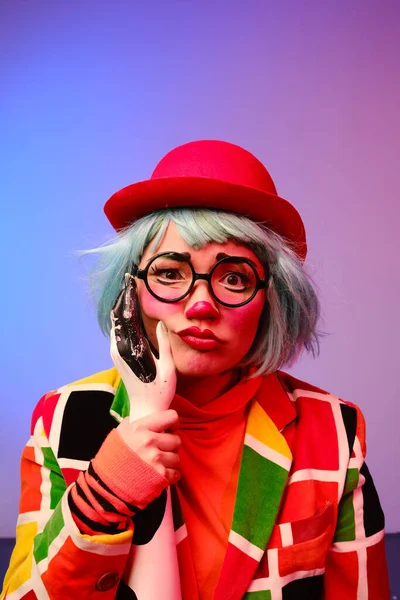 Close-up portrait of a clown girl with make-up, blue hair, a red hat, a colored checkered jacket and glasses.