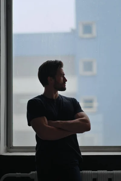 Retrato de um jovem homem elegante com barba e cabelo escuro em uma camiseta preta fica perto da janela — Fotografia de Stock