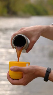 Erkek gezgin, nehrin arka planına termostan sıcak bir içecek dolduruyor.