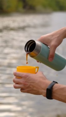 Erkek gezgin, nehrin arka planına termostan sıcak bir içecek dolduruyor.