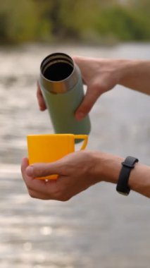 Erkek gezgin, nehrin arka planına termostan sıcak bir içecek dolduruyor.