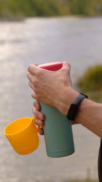 Männlicher Reisender öffnet eine Thermoskanne, um Tee in einen gelben Plastikbecher auf dem Hintergrund des Flusses zu gießen. — Stockvideo