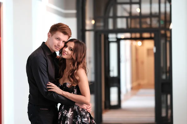 Jovem casal bonito - uma menina em um vestido magnífico e um cara em uma camisa preta e calças caminham ao longo do edifício de tijolos isóico — Fotografia de Stock