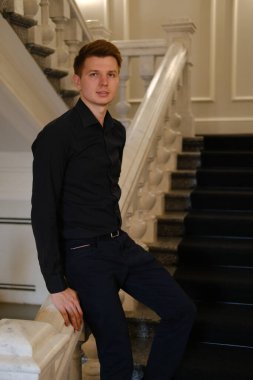 Siyah gömlekli ve siyah pantolonlu genç bir adam tarihi bir binanın beyaz merdivenlerinde oturuyor.