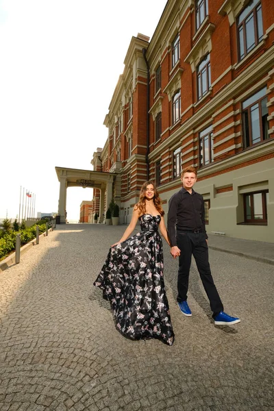 Młoda piękna para - dziewczyna w wspaniałej sukience i facet w czarnej koszuli i spodniach spacerują po izoicznym budynku z cegły — Zdjęcie stockowe