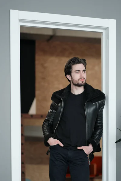 Un hombre joven y elegante con barba y cabello oscuro en una chaqueta de cuero negro con cuello de piel sintética — Foto de Stock