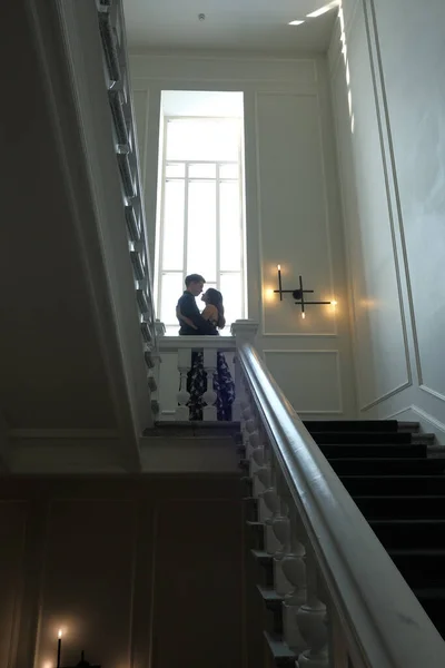 Joven pareja hermosa - una chica y un chico están de pie o sentados en una escalera blanca dentro de un hermoso edificio grande . — Foto de Stock