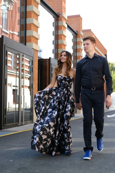 Jovem casal bonito - uma menina em um vestido magnífico e um cara em uma camisa preta e calças caminham ao longo do edifício de tijolos isóico — Fotografia de Stock