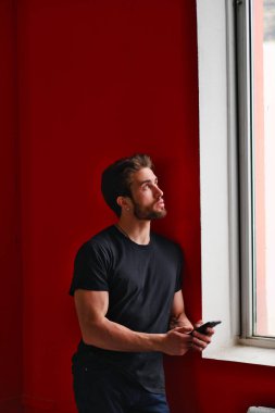 Siyah tişörtlü, sakallı, koyu renk saçlı genç bir adamın portresi pencerenin yanında duruyor. 