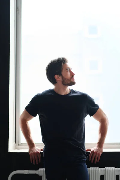 Retrato de um jovem homem elegante com barba e cabelo escuro em uma camiseta preta fica perto da janela — Fotografia de Stock