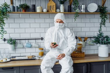 Koruyucu beyaz takım elbiseli, kapüşonlu ve maske takan Avrupalı bir adamın portresi evden telefonla çalışıyor ve önemli bir işi çözüyor. Koronavirüs salgını nedeniyle karantinaya alınmış ve izole edilmiş.
