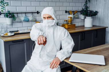 Viral beyaz takım elbiseli ve yüz maskeli bir adam evde bilgisayarla oturuyor ve kamerayı parmağıyla işaret ediyor. Koronavirüs salgını ve karantina yüzünden izole edilmiş.