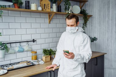 Viral koruyucu kostüm ve yüz maskesi takan Avrupalı bir adamın portresi mutfakta duruyor ve koronavirüs salgını sırasında telefonu arıyor. Güvenlik için karantina sırasında evden çalışıyor.