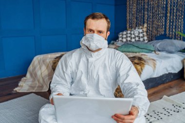 Beyaz viral koruyucu kostümlü beyaz adam portresi ve yüz maskesi yatağın yanında oturuyor ve Coronavirus yüzünden dizüstü bilgisayarla çalışıyor. Covid-19 karantinasından dolayı evde izole edilmiş.