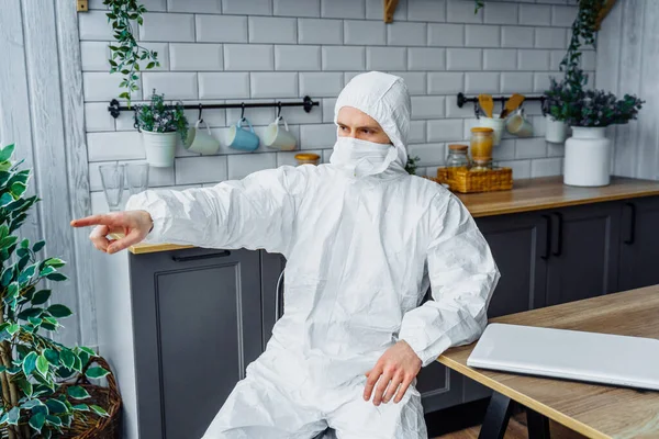 Viral beyaz takım elbiseli ve yüz maskeli bir adam evde bilgisayarla oturuyor ve pencereyi parmağıyla işaret ediyor. Koronavirüs salgını ve karantina yüzünden izole edilmiş.