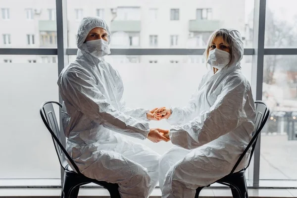 Doctors sitting on the chairs, holding hands in front of the window in hospital. They love and support each other, wearing protective white suits and face masks due to epidemic of coronavirus covid-19