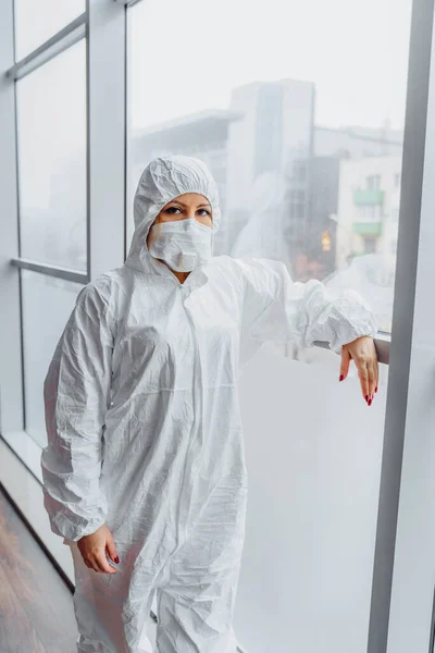 Sad doctor standing near window in hospital in protective white suit and face mask because of coronavirus epidemy. Portrait of upset, tired and worried doctor woman thinking of covid-19 pandemic