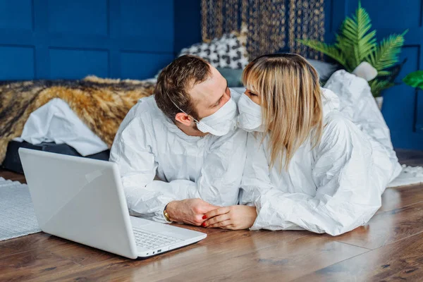 Be safe and stay at home! Doctors lying on the floor wearing face masks against coronavirus and kissing. People in protective suits isolated due to epidemic of covid-19 and working on laptop