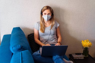 Koruyucu maske takan çekici sarışın kız evde uzaktan kumandalı bir bilgisayarla çok çalışıyor. Dünya koronavirüs salgınından dolayı izole edilmiş. Covid-19 karantina döneminde.